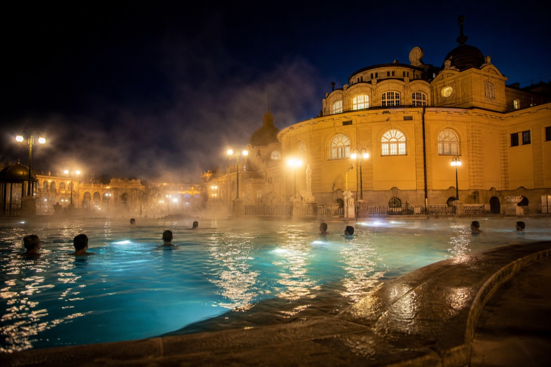 Szechenyi outdoor pool at night with lit yellow facade and steam rising into the dark sky