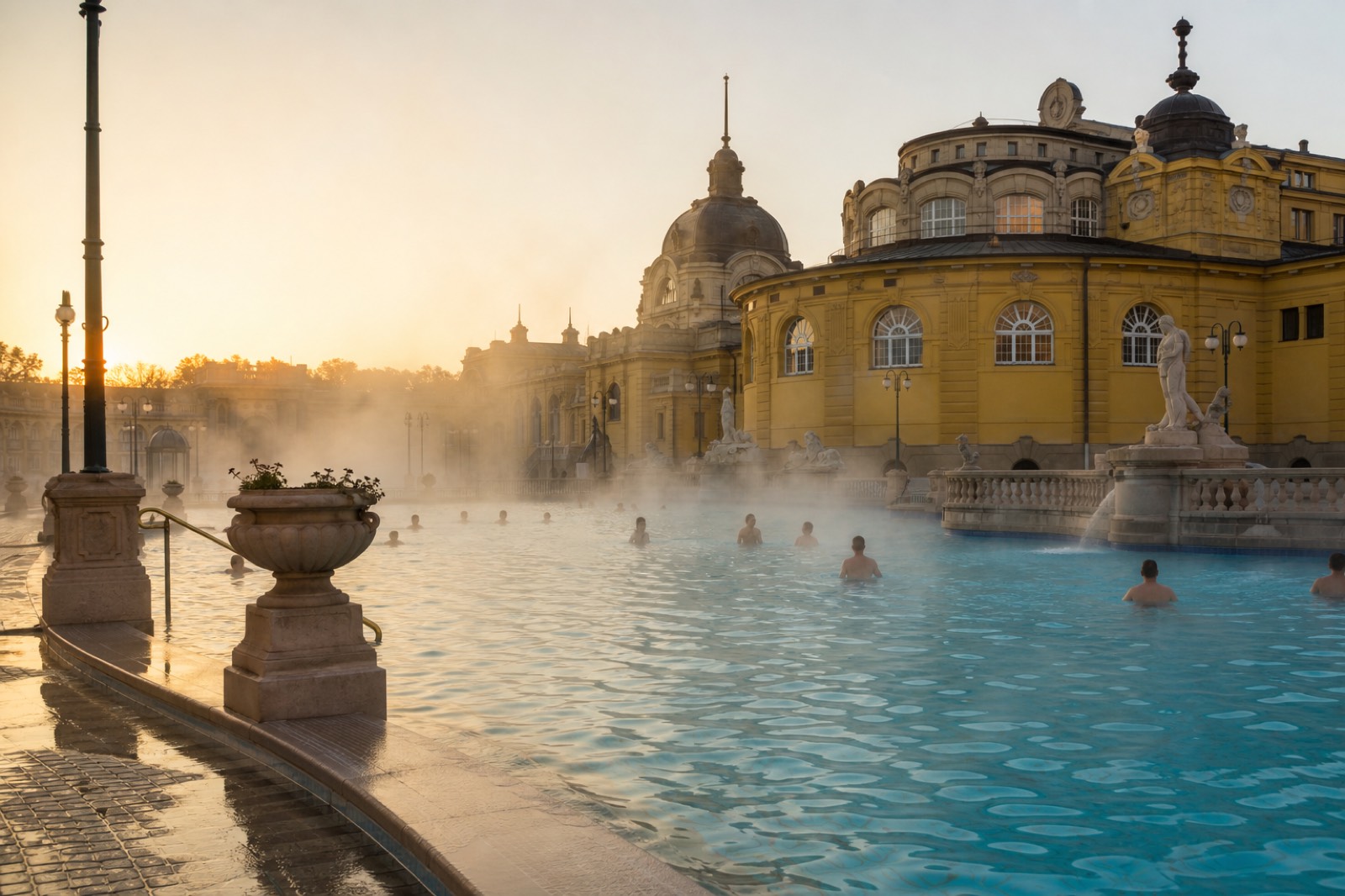 Steam rising off the outdoor thermal pool at Szechenyi bath at sunrise with the yellow facade behind it