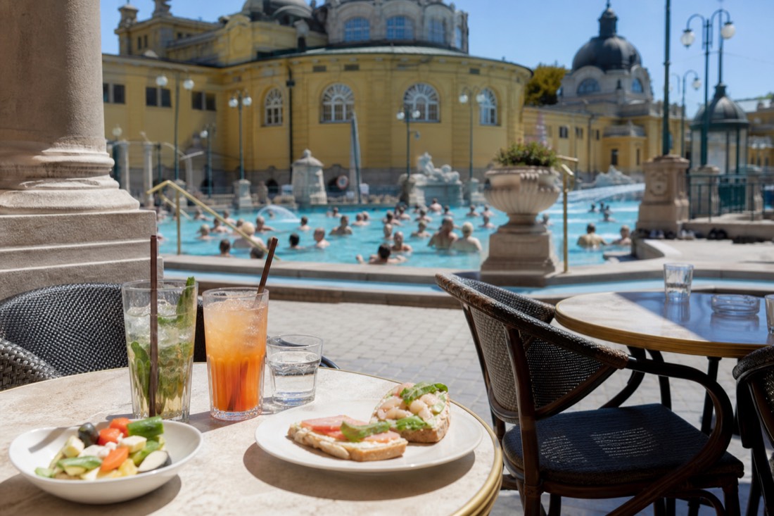 Poolside terrace tables with drinks overlooking an outdoor thermal pool in Budapest