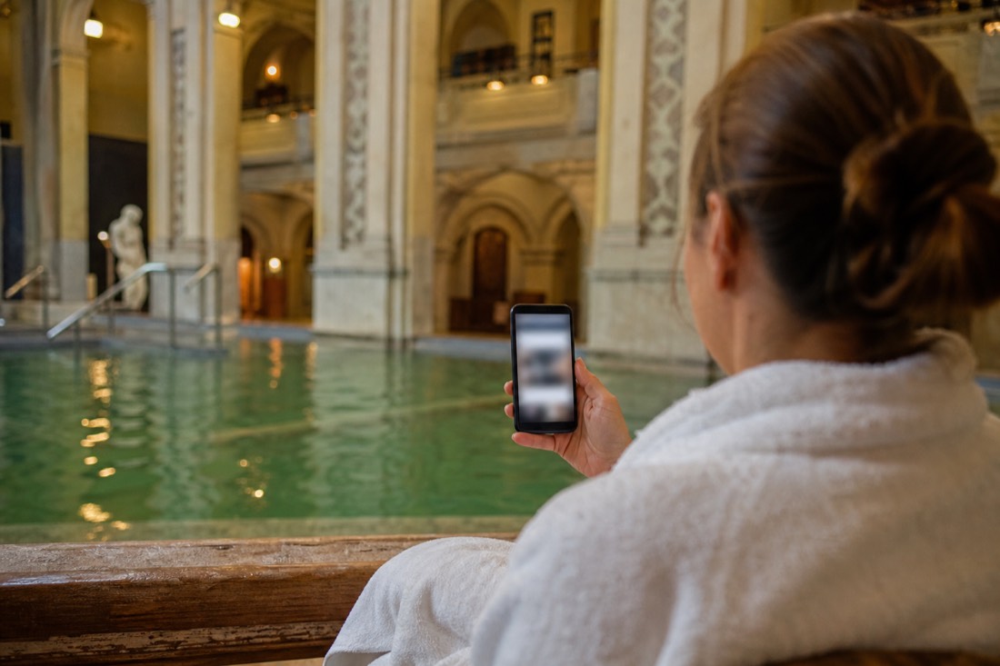 Visitor reading information on a phone beside an indoor thermal pool in Budapest