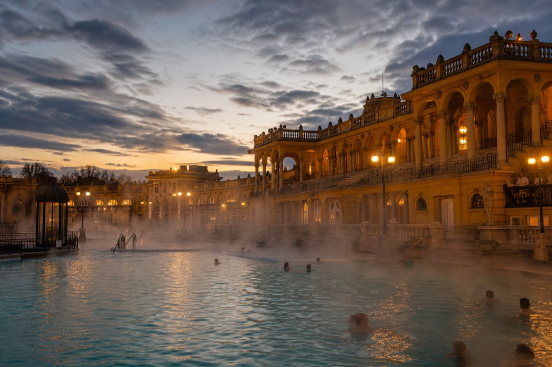 Steam rising off the outdoor thermal pool at a grand yellow Budapest bath in the evening