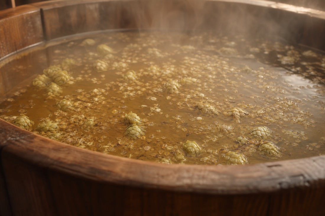 Amber-tinted water steaming in a wooden beer spa tub with hops floating on the surface