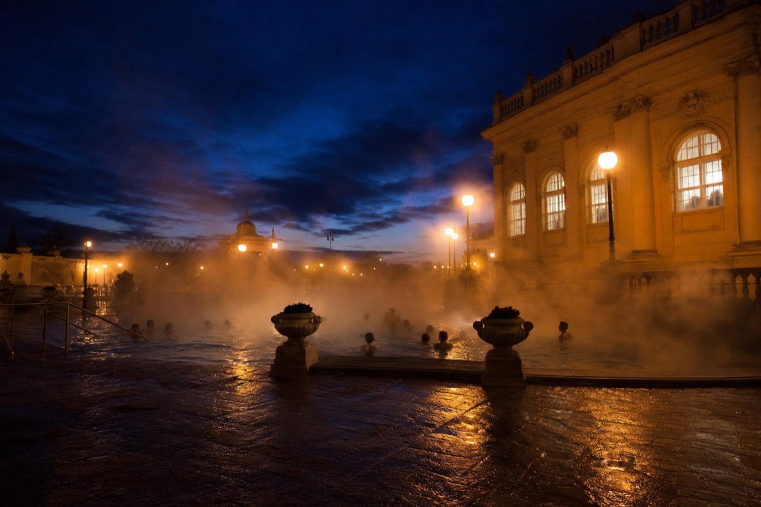Outdoor thermal pool glowing at dusk with steam and warm lights on a baroque bathhouse facade