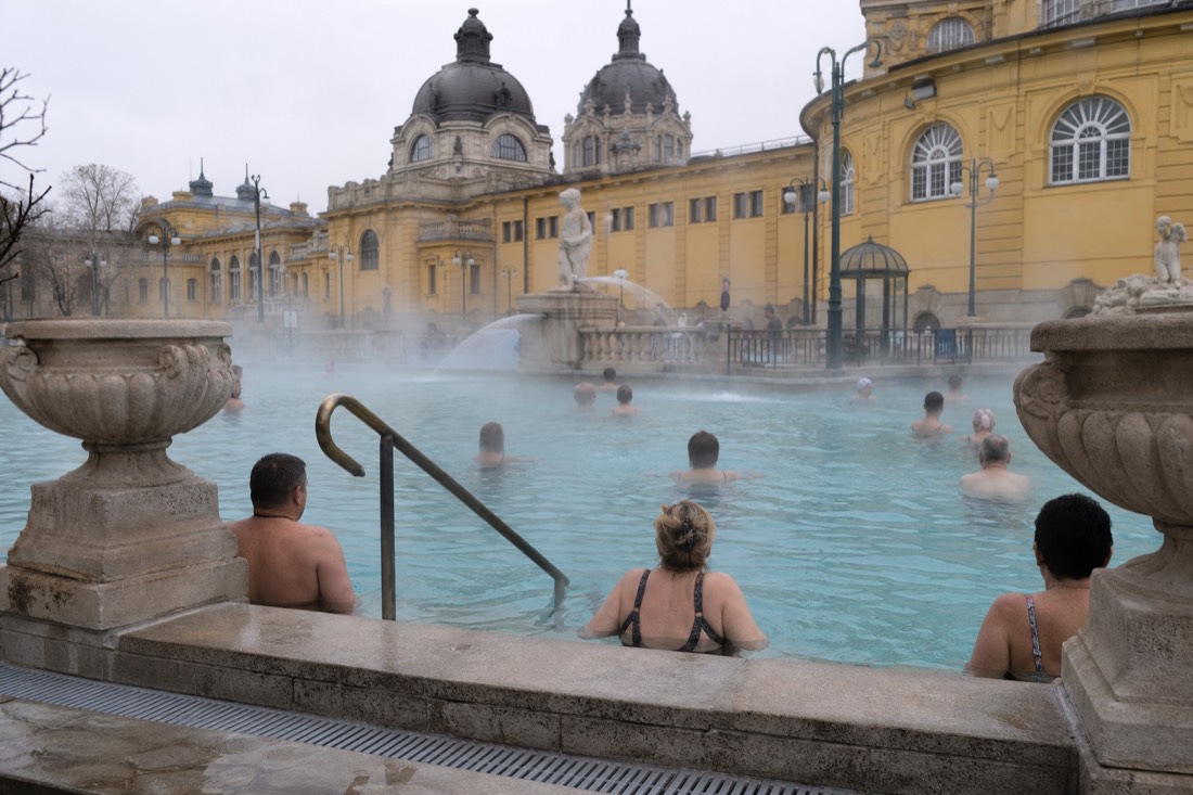 Bathers in ordinary swimsuits relaxing along the edge of a steaming outdoor thermal pool in Budapest