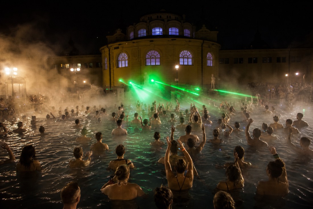 Crowd of guests seen from behind in a steaming outdoor pool under lasers at a Budapest night spa event