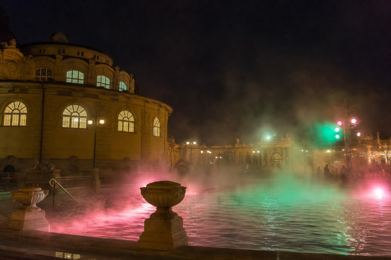 Colored party lights and steam rising over the outdoor pool of a grand Budapest thermal bath at night