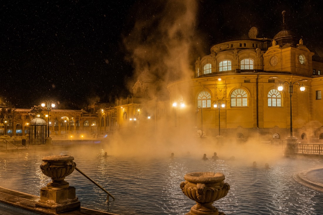 Yellow baroque bath facade lit at night with steam rising from the outdoor pool into falling snow