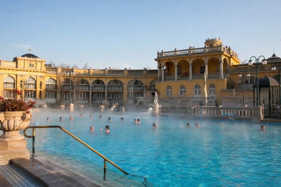Turquoise outdoor thermal pool framed by a neo-baroque yellow facade on a bright Budapest morning