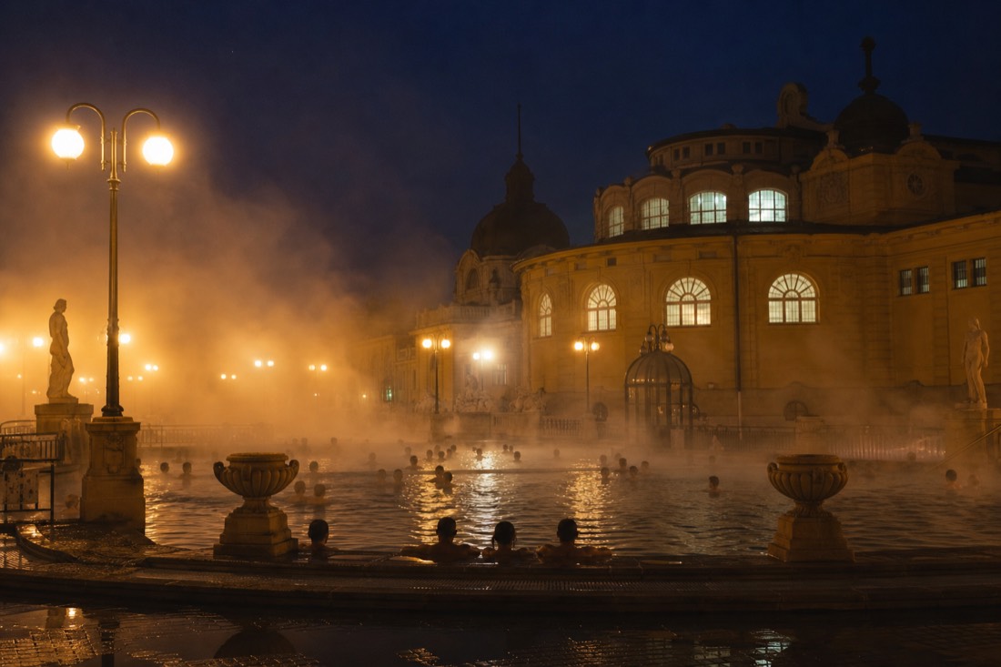 Steam rising from an outdoor thermal pool at night with bathers relaxing under warm lights