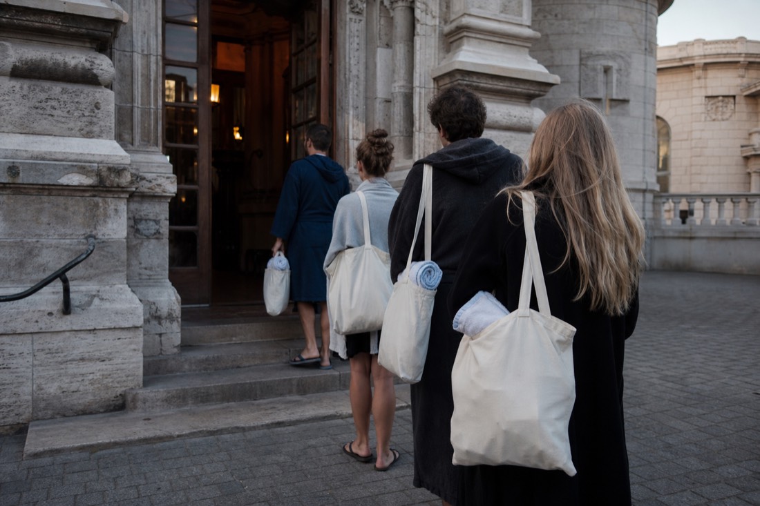 Early morning queue of visitors with tote bags outside a neo-baroque Budapest bathhouse entrance