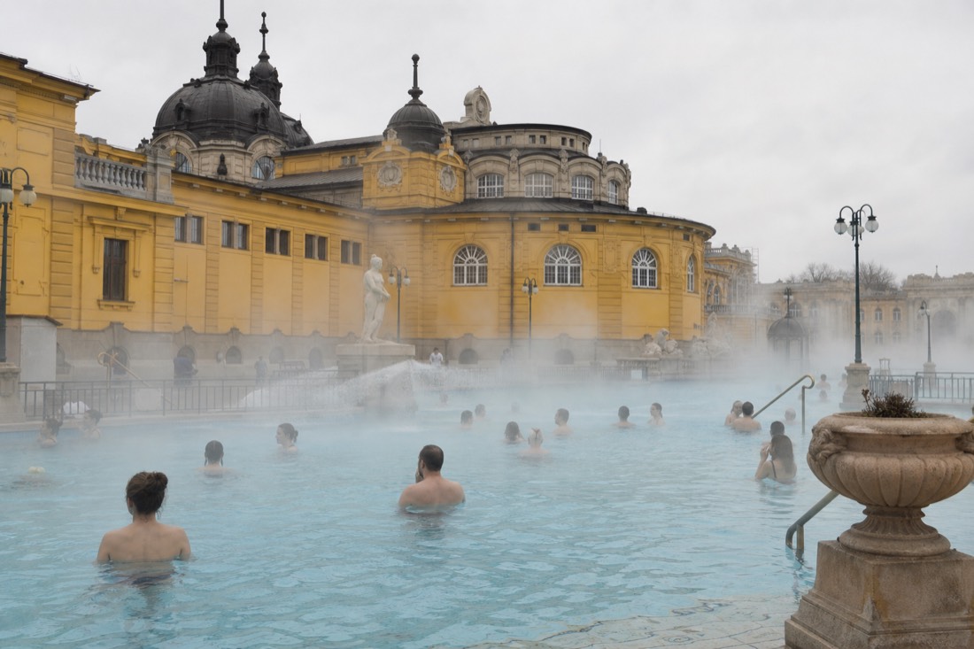 Bathers relaxing in a steaming outdoor thermal pool beside ornate yellow bathhouse walls