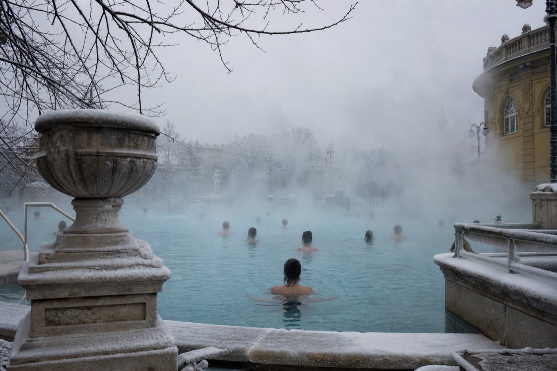 Bathers in a steaming outdoor thermal pool in Budapest with winter sky overhead