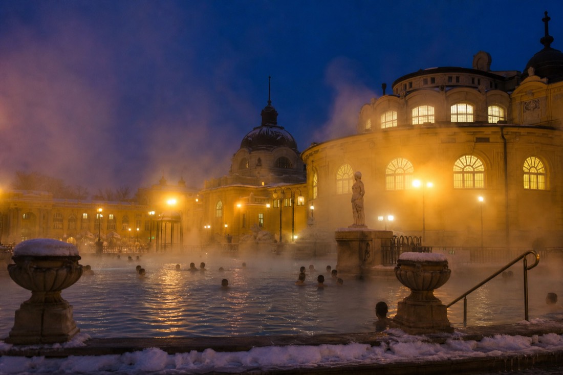 Evening steam over the outdoor pools of a grand yellow bath courtyard in Budapest under a winter sky