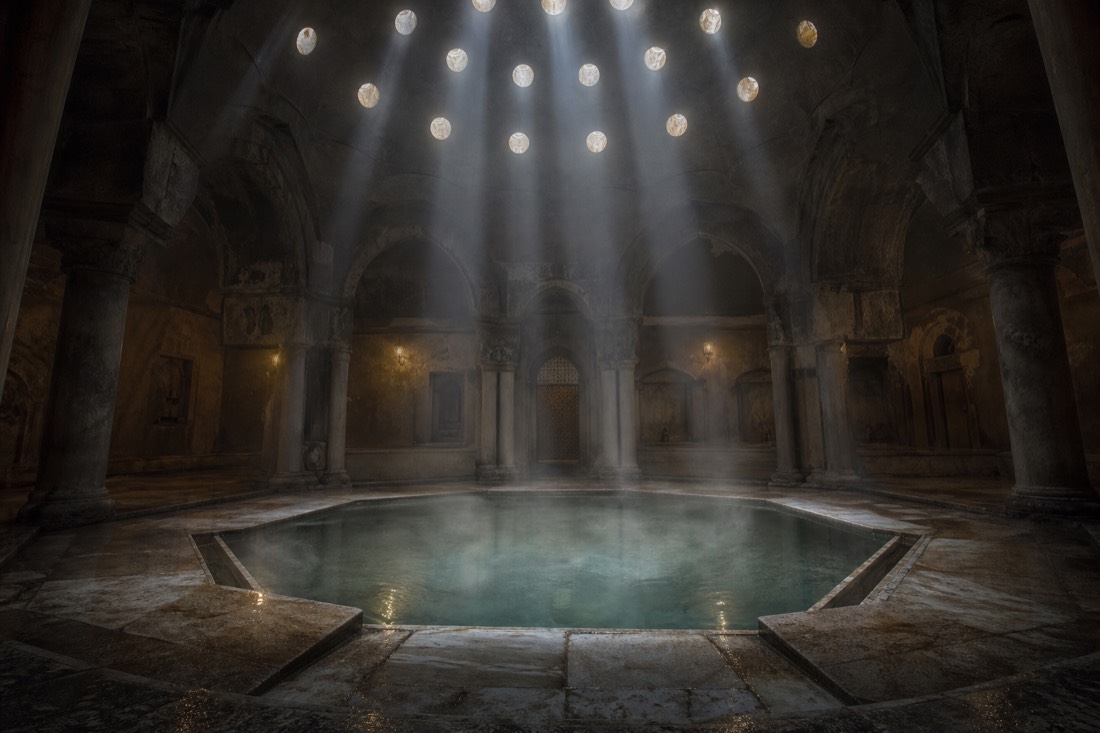 Shafts of light falling through a domed stone ceiling onto an octagonal thermal pool in an Ottoman-era bath