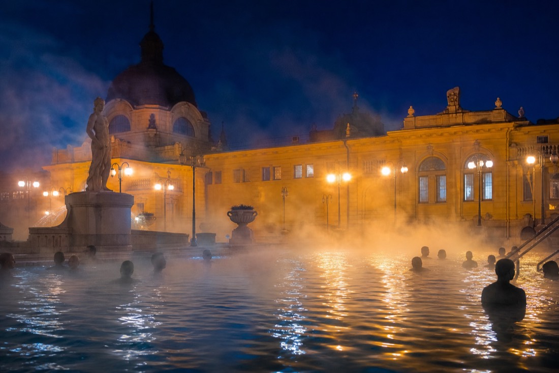 Evening steam rising from an outdoor thermal pool in Budapest with warm lights on the surrounding building