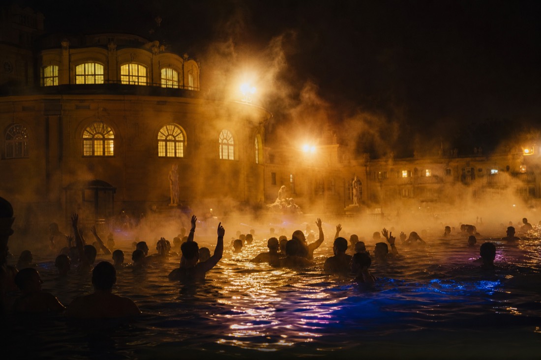 Steam glowing over an outdoor bath pool lit up at night with the silhouettes of a crowd in the water