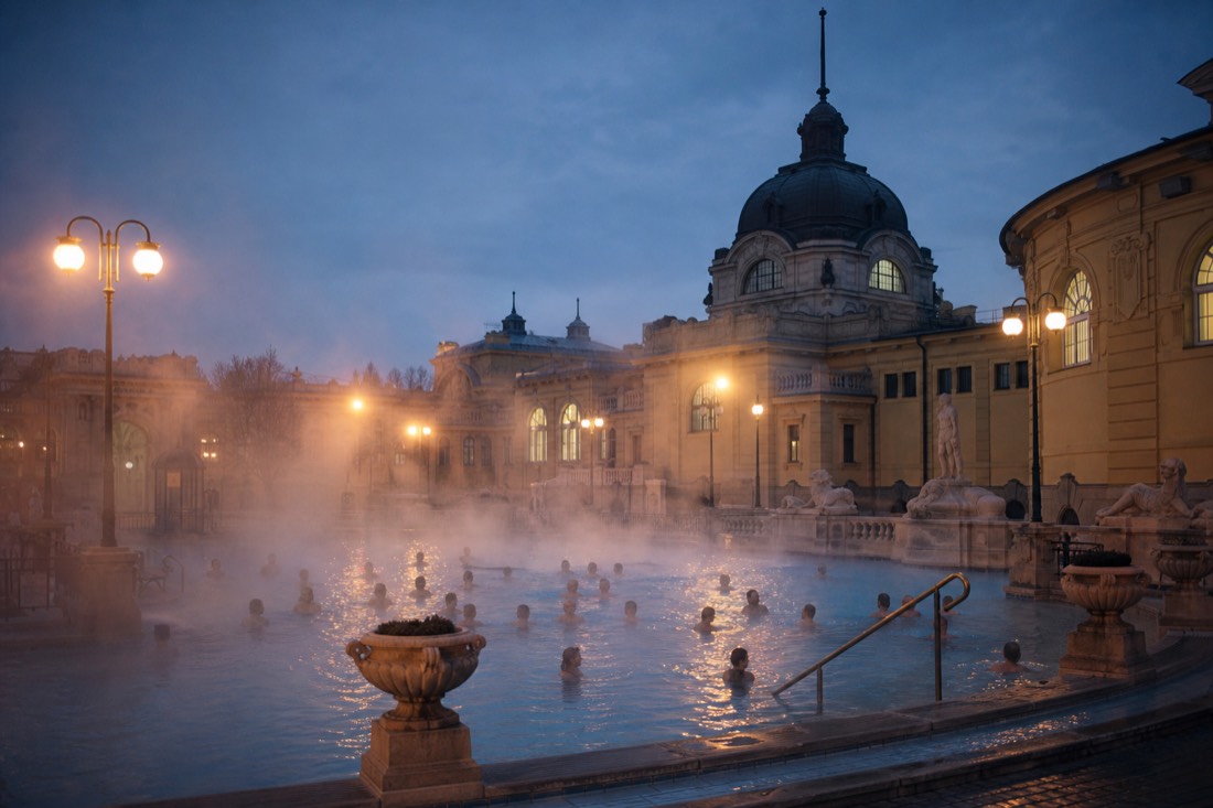 Steam rising over swimmers wearing swimsuits in a historic outdoor thermal pool at dusk in Budapest