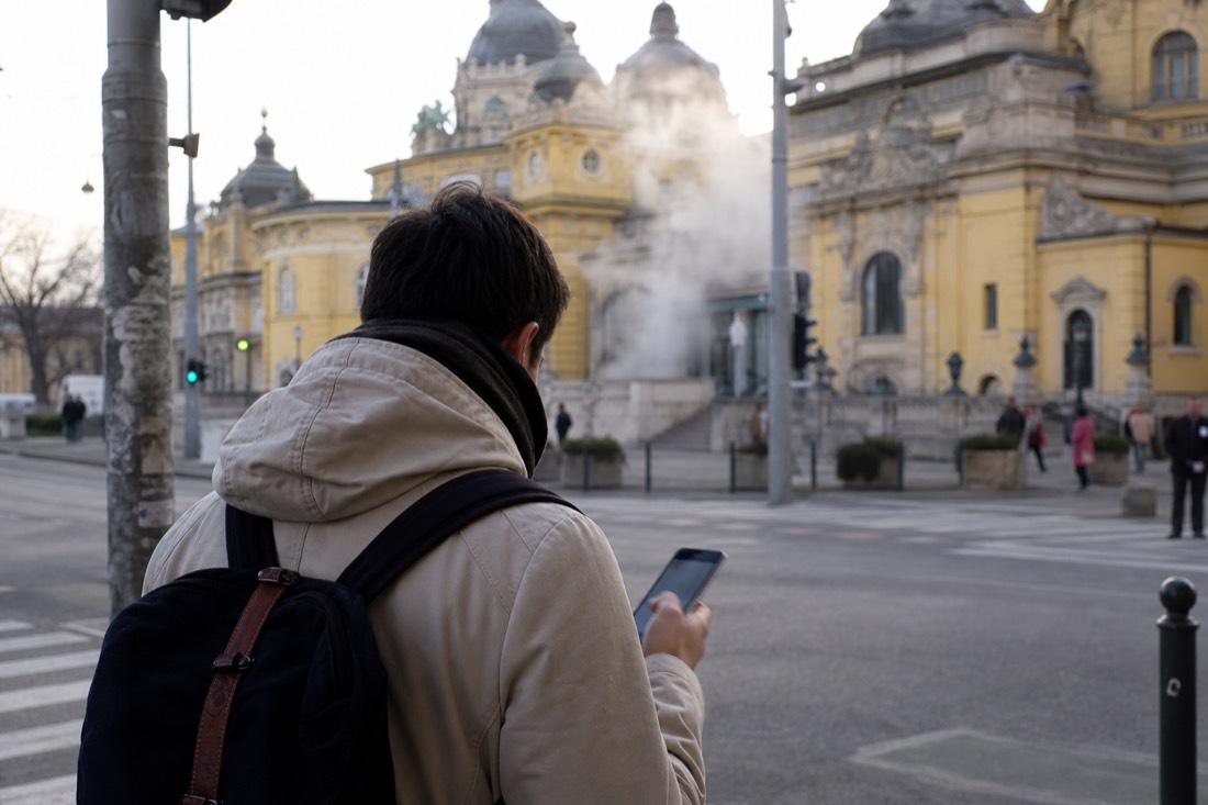 Traveller reading a phone outside a Budapest bathhouse while steam drifts from the building behind